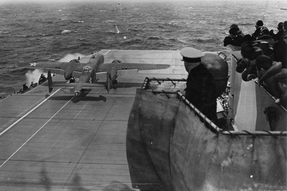 In this iconic photo, Jimmy Doolittle’s aircraft is seen taking off first from the deck of the USS Hornet on the morning of April 18, 1942, at 08:20. As the commander of the mission, Doolittle led the way. His B-25 Mitchell bomber, heavily loaded and modified for extended range, had only about 140 meters of takeoff space on the ship. The long white lines painted on the deck served as visual guides to help the pilots maintain a straight path during their very short takeoff roll. All sixteen B-25s participating in the Doolittle Raid launched from the USS Hornet and successfully became airborne without any significant mechanical issues during takeoff. Despite the short runway and the rough conditions at sea, each aircraft managed to lift off—an impressive feat, given that this had never been attempted before with medium bombers. The idea of launching medium bombers from an aircraft carrier had been considered impossible. Yet, through intense training, specially modified aircraft, and favorable wind conditions, these highly skilled crews accomplished a stunt previously unknown in military aviation. Naval officers and crew members of the USS Hornet watched tensely as the mission began to unfold.